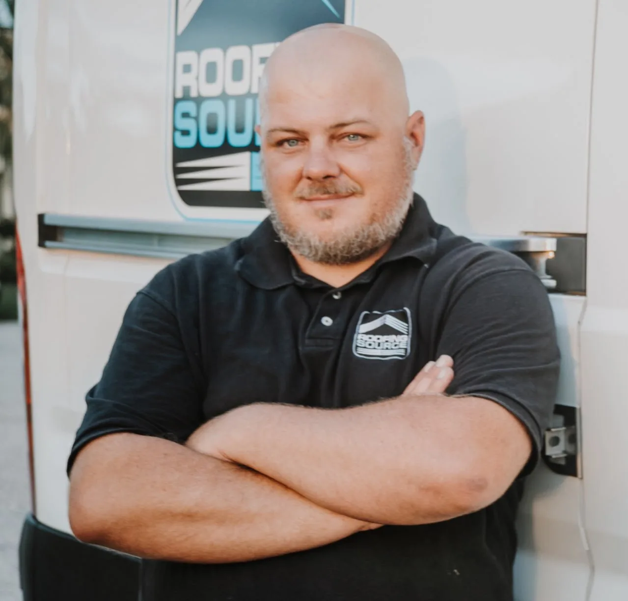 A RoofingSource teammate in a black company polo, arms folded, in front of a company van