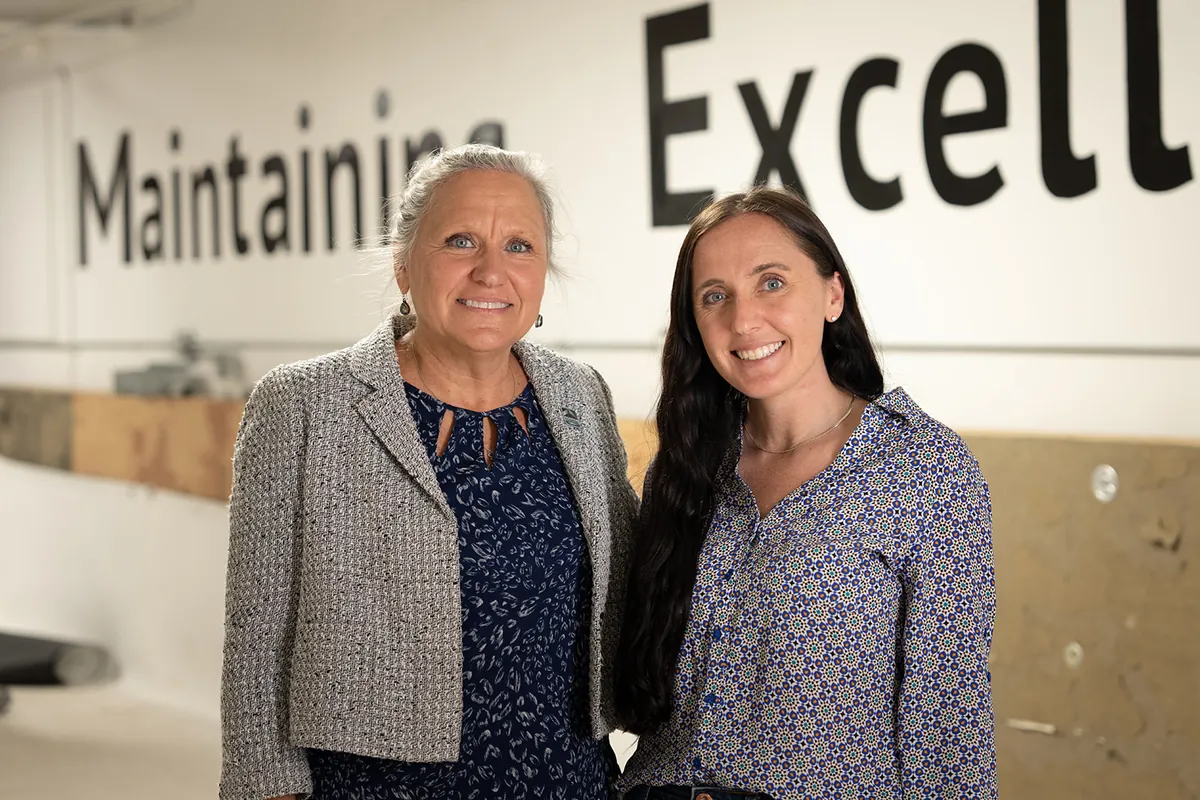 Two teammates in front of the Maintaining Excellence wall at headquarters
