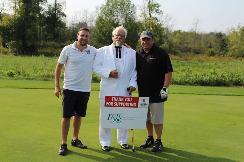Three teammates on a golf course beside a charity sponsorship sign