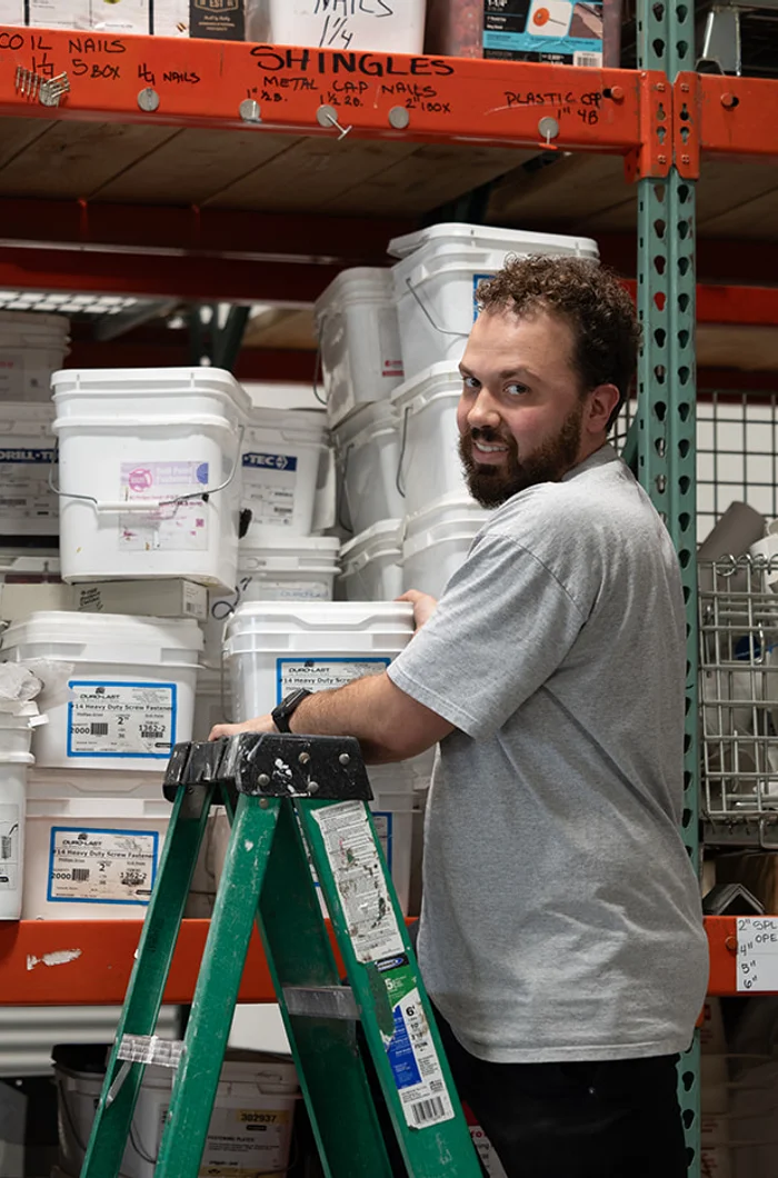 A smiling RoofingSource teammate beside a ladder in the headquarters supply warehouse