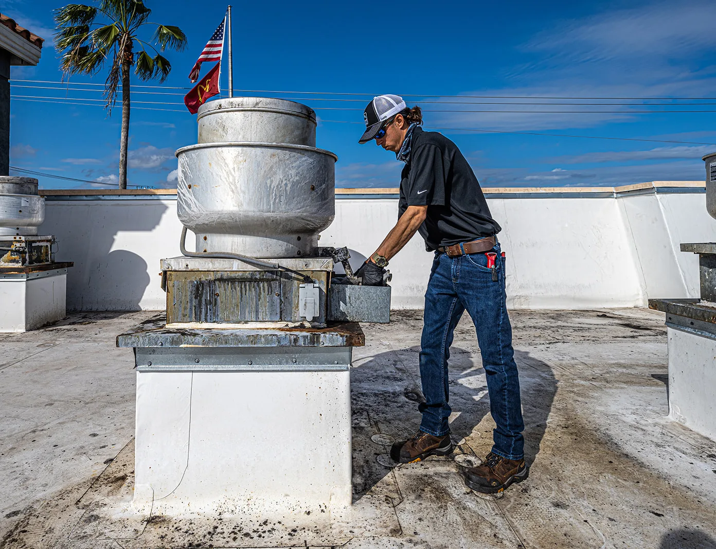 A RoofingSource teammate in a cap working at the base of a rooftop exhaust fan