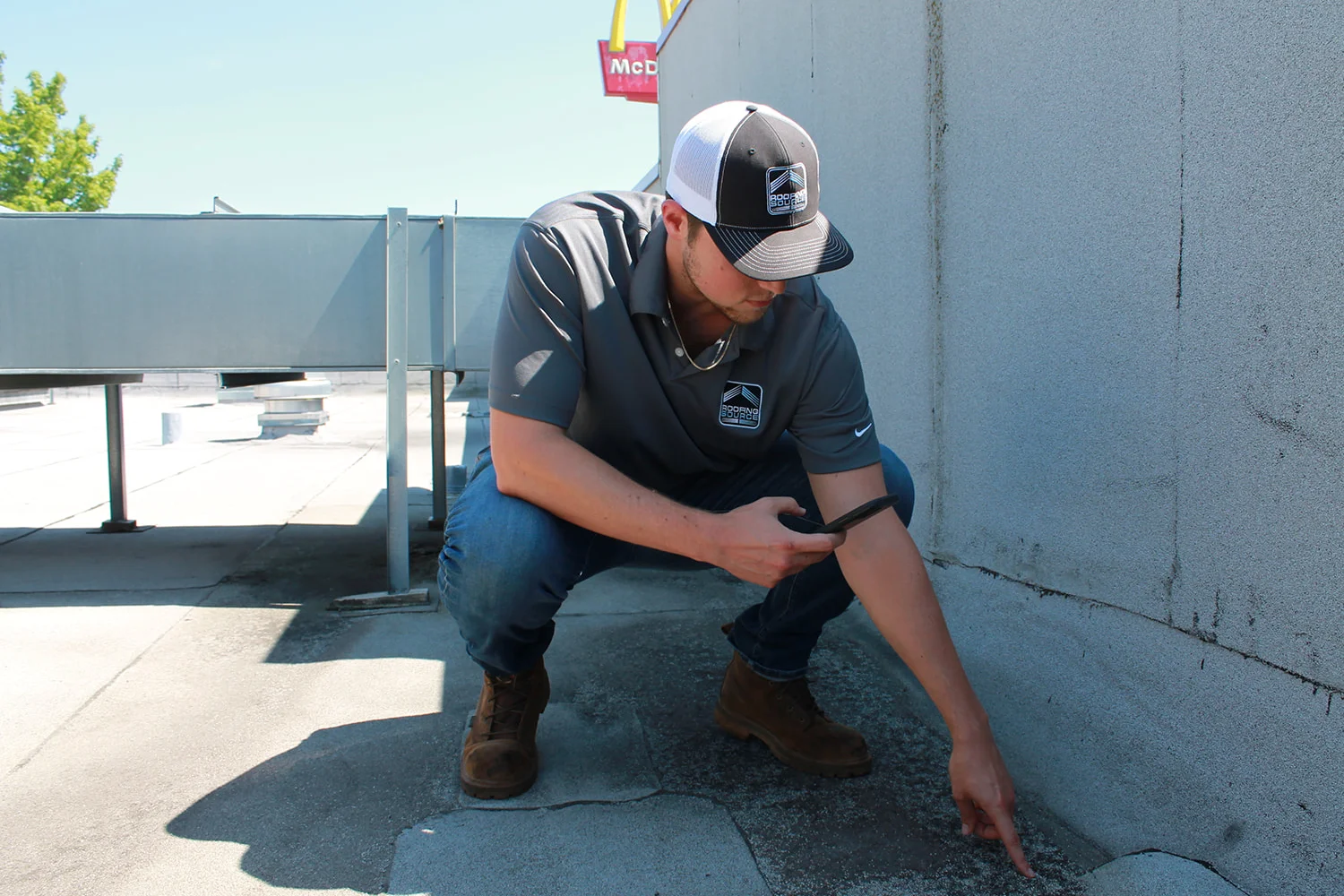 A Field Advisor crouched at a parapet wall, phone in hand, pointing out a defect at its base